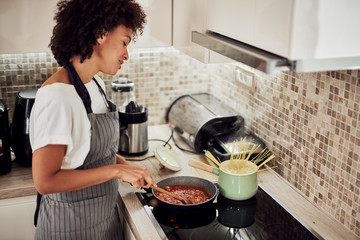 Mixed race woman in apron standing next to stove in domestic kitchen and making pasta and sauce.