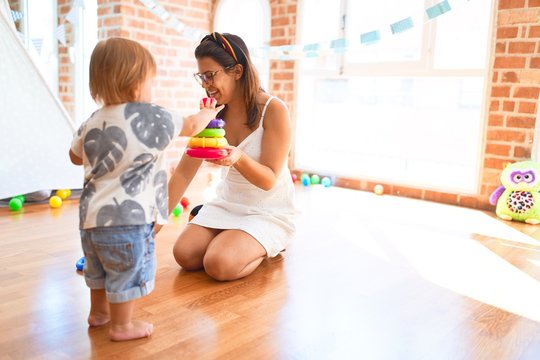Beautiful teacher and toddler building pyramid using hoops around lots of toys at kindergarten