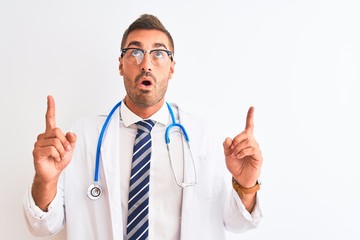Young handsome doctor man wearing stethoscope over isolated background amazed and surprised looking up and pointing with fingers and raised arms.