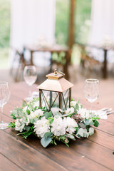 The composition of flowers and greenery, golden lantern in rustic style, standing on served table in the area of wedding party outdoors