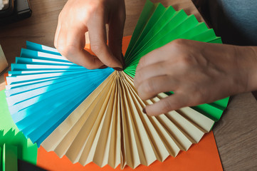 In the foreground female hands glue origami fans, sheets of colored paper, scissors on a wooden...
