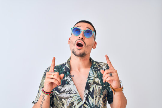 Young handsome man wearing Hawaiian sumer shirt and blue sunglasses over isolated background amazed and surprised looking up and pointing with fingers and raised arms.