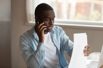 Focused young black man holding documents, calling to financial advisor.