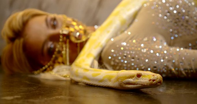A close-up portrait of a blonde girl, she is lying on the floor, she has a beautiful mask of jewelry on her face, she herself is dressed in a tight-fitting suit with sequins, a snake is lying on her.