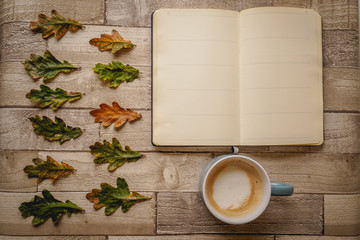 Top view of an open notebook and a cappuccino mug with green and orange leaves on a wooden board. Landscape format.