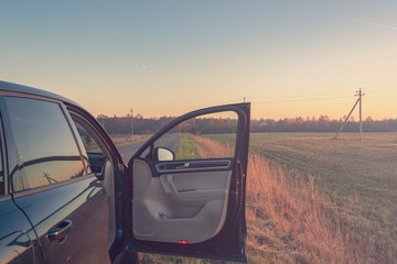 The open door of the car standing on the side of the road against the backdrop of the autumn landscape at sunset