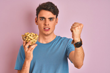 Teenager boy holding a bunch of peanunts over isolated pink background annoyed and frustrated shouting with anger, crazy and yelling with raised hand, anger concept