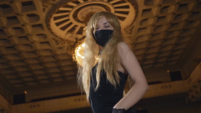 Sexy girl in black dress and black dust mask posing in front of an ancient ceiling in the concert hall.