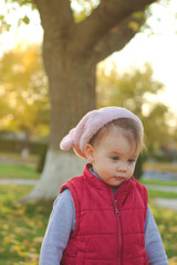 A small child in a fluffy pink hat and red vest is running in the autumn park. Beautiful fall sunny day outdoors.