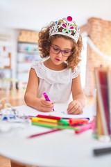 Beautiful toddler wearing glasses and princess crown sitting drawing using paper and marker pen at kindergarten