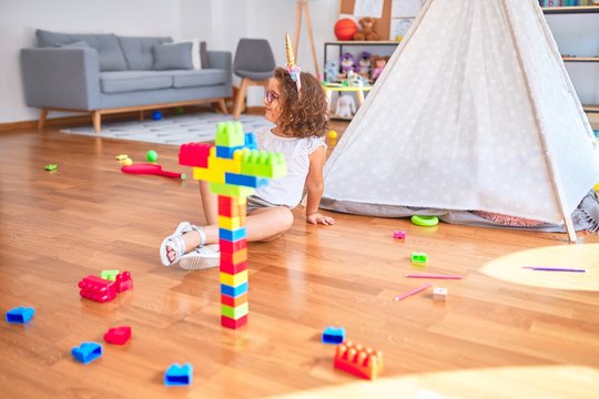 Beautiful toddler wearing glasses and unicorn diadem sitting playing with building blocks smiling at kindergarten