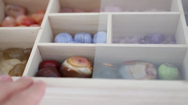 Polychrome jasper, blue agate, amethyst, citrin,  rose quartz lie in a wooden box with a transparent lid. Female hand opens the box cover