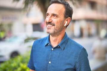 Middle age handsome businessman standing on the street listening to music using earphones smiling
