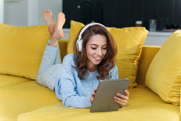 Exuberant girl uses tablet pc while laying on the comfortable yellow sofa and enjoying nice melodies with earphones.