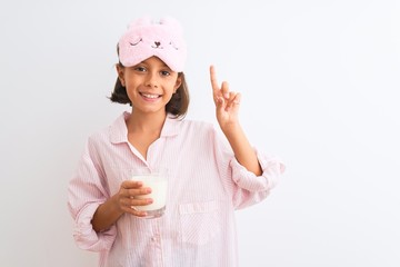 Child girl wearing sleep mask and pajama drinking glass of milk over isolated white background surprised with an idea or question pointing finger with happy face, number one