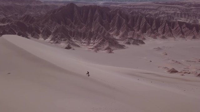 Man climbing upholding a sandboard over great Sand Dunes of the Dead Valley, Atacama desert. Aerial view, 4k