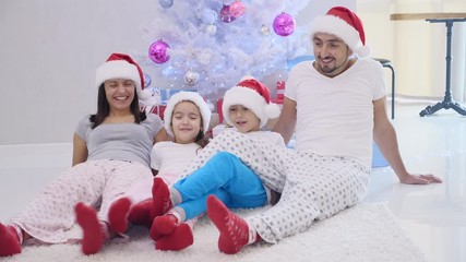 Joyful father mother sister brother are ready for christmas, sitting on the carpet near fir-tree, dressed in santa hats and red socks, playing around.