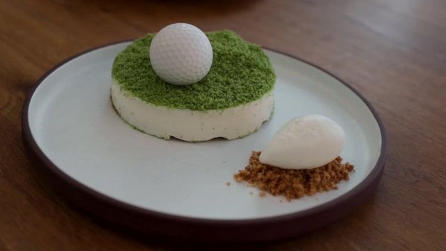 Gourmet dessert, decorated in concept of a golf ball on grass, served on a flat round stoneware plate with a tall rim, set on a wood dinner table.