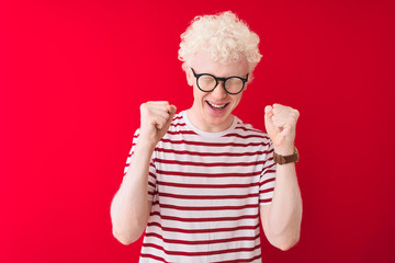 Young albino blond man wearing striped t-shirt and glasses over isolated red background excited for success with arms raised and eyes closed celebrating victory smiling. Winner concept.