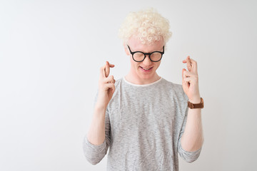 Young albino blond man wearing striped t-shirt and glasses over isolated white background gesturing finger crossed smiling with hope and eyes closed. Luck and superstitious concept.
