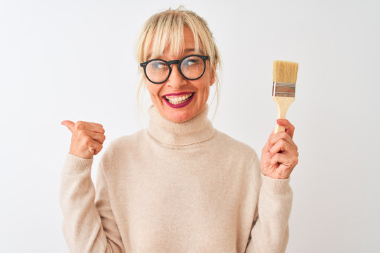 Middle age painter woman holding paint brush standing over isolated white background pointing and showing with thumb up to the side with happy face smiling