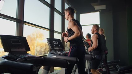 Two men running on a treadmill in the background of a large window in the fitness room. Young men running in the fitness room. Aerobic workouts in the gym - Powered by Adobe