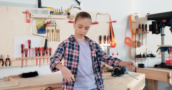 Young girl holding measuring tape, standing near desk in wood workshop