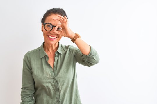 Middle age woman wearing green shirt and glasses standing over isolated white background doing ok gesture with hand smiling, eye looking through fingers with happy face.