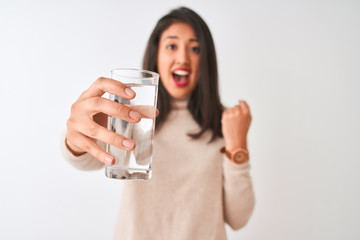 Fototapeta premium Young beautiful chinese woman holding glass of water standing over isolated white background screaming proud and celebrating victory and success very excited, cheering emotion