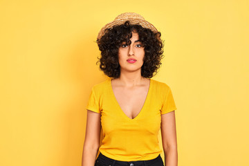 Young arab woman with curly hair wearing t-shirt and hat over isolated yellow background with serious expression on face. Simple and natural looking at the camera.