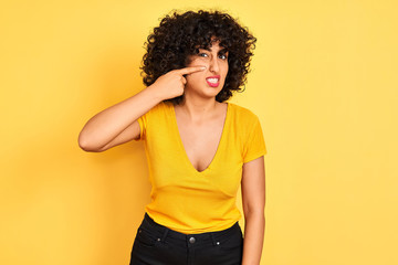 Young arab woman with curly hair wearing t-shirt standing over isolated yellow background pointing unhappy to pimple on forehead, ugly infection of blackhead. Acne and skin problem
