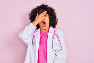 Young arab doctor woman with curly hair wearing stethoscope over isolated pink background peeking in shock covering face and eyes with hand, looking through fingers with embarrassed expression.