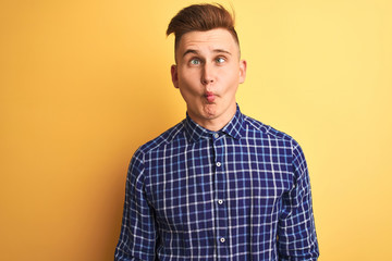 Young handsome man wearing casual shirt standing over isolated yellow background making fish face with lips, crazy and comical gesture. Funny expression.