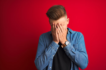 Young handsome man wearing denim shirt standing over isolated red background with sad expression covering face with hands while crying. Depression concept.