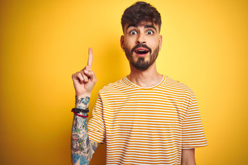 Young man with tattoo wearing striped t-shirt standing over isolated yellow background pointing...