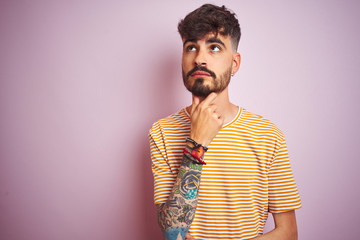 Young man with tattoo wearing yellow striped t-shirt standing over isolated pink background with hand on chin thinking about question, pensive expression. Smiling with thoughtful face. Doubt concept.