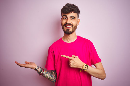 Young man with tattoo wearing t-shirt standing over isolated pink background amazed and smiling to the camera while presenting with hand and pointing with finger.