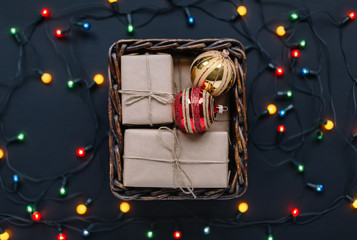 Wooden basket with few presents, yellow and red fir balls. Gift boxes wrapped in kraft paper. Around basket shine colorful lights of garland. Merry Christmas mood. Happy New Year preparation. Close up
