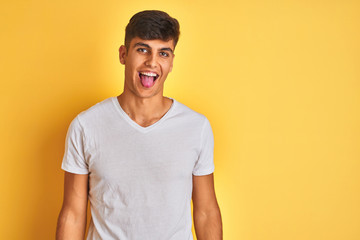 Fototapeta premium Young indian man wearing white t-shirt standing over isolated yellow background sticking tongue out happy with funny expression. Emotion concept.