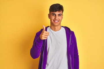 Young indian man wearing purple sweatshirt standing over isolated yellow background doing happy thumbs up gesture with hand. Approving expression looking at the camera showing success.