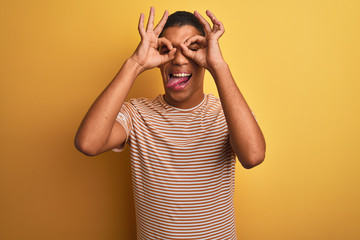 Young handsome arab man wearing striped t-shirt standing over isolated yellow background doing ok gesture like binoculars sticking tongue out, eyes looking through fingers. Crazy expression.