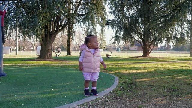 Dolly out slow motion shot as pretty mixed race black baby infant smiles and feels life around her in the sunny morning park dressed in pink stuffed hoodie.