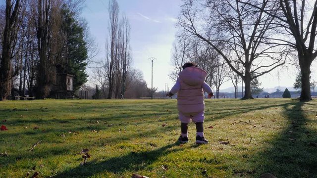 Slow motion shot of a baby girl infant mixed race dressed in pink with hoodie walking off in a park in stumbling movements as she just learned to walk.