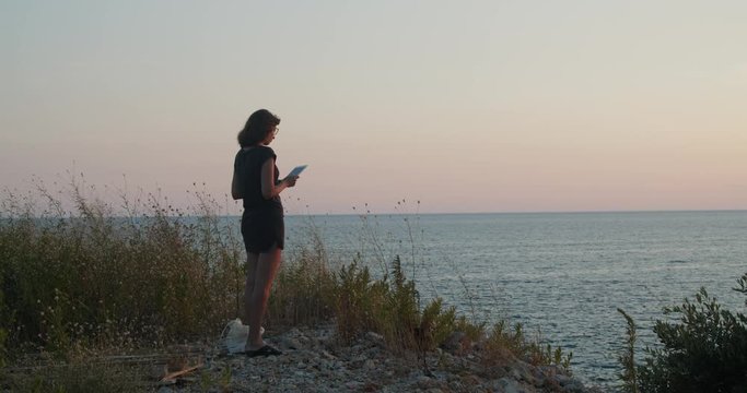 Young woman standing on a cliff by the sea on sunset, takes a picture with tablet computer , wide shot