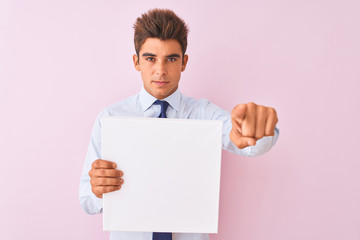 Young handsome businessman holding banner standing over isolated pink background pointing with finger to the camera and to you, hand sign, positive and confident gesture from the front