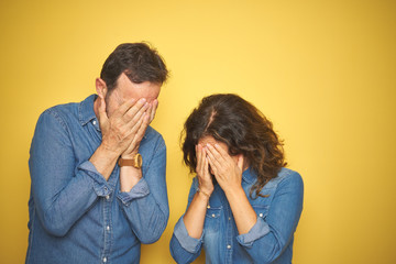 Beautiful middle age couple together standing over isolated yellow background with sad expression covering face with hands while crying. Depression concept.