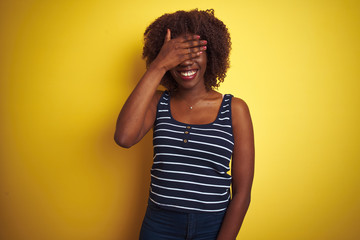 Young african afro woman wearing striped t-shirt over isolated yellow background smiling and laughing with hand on face covering eyes for surprise. Blind concept.