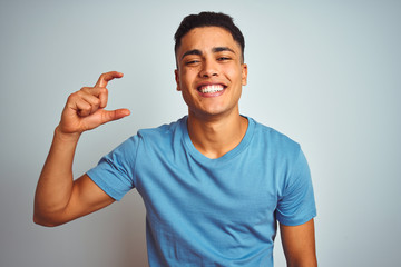 Young brazilian man wearing blue t-shirt standing over isolated white background smiling and confident gesturing with hand doing small size sign with fingers looking and the camera. Measure concept.