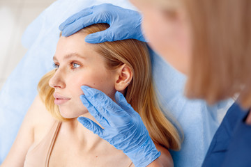 Cosmetology Service. Young woman at beauty clinic lying while doctor in gloves touching cheek...
