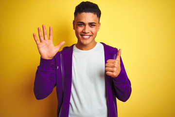 Young brazilian man wearing purple sweatshirt standing over isolated yellow background showing and pointing up with fingers number six while smiling confident and happy.